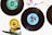 Vinyl records are splayed out on a white surface, with labels in different colors, such as green and blue. A skateboard wheel and truck, positioned in the lower left corner, add an eclectic touch to the arrangement. The top portion of a record sleeve is visible in the corner, featuring printed text and abstract artwork.