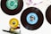 Vinyl records are splayed out on a white surface, with labels in different colors, such as green and blue. A skateboard wheel and truck, positioned in the lower left corner, add an eclectic touch to the arrangement. The top portion of a record sleeve is visible in the corner, featuring printed text and abstract artwork.