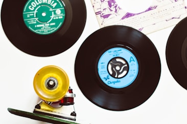 Vinyl records are splayed out on a white surface, with labels in different colors, such as green and blue. A skateboard wheel and truck, positioned in the lower left corner, add an eclectic touch to the arrangement. The top portion of a record sleeve is visible in the corner, featuring printed text and abstract artwork.