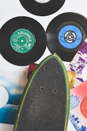 A collection of vintage vinyl records with colorful labels is arranged alongside a skateboard. The records include labels such as Columbia and Chrysalis, showcasing various designs with colors like green and blue. The skateboard, with a black grip and green edges, is positioned diagonally, adding to the retro aesthetic.