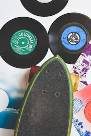 A collection of vintage vinyl records with colorful labels is arranged alongside a skateboard. The records include labels such as Columbia and Chrysalis, showcasing various designs with colors like green and blue. The skateboard, with a black grip and green edges, is positioned diagonally, adding to the retro aesthetic.