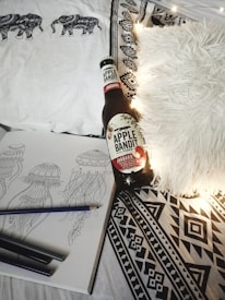 A cozy arrangement featuring a bottle of Apple Bandit Cider on a bed with patterned sheets. There is a sketchbook open to drawings of jellyfish with colored pencils beside it. A fluffy white pillow and a string of warm lights add to the comfortable ambiance.