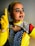 A person is dressed in a pop art-inspired costume featuring bright colors and dotted face paint to mimic a comic book character. The face has red dots and bold black lines emphasizing facial features. The outfit includes a yellow top, gloves, and a cape or scarf with polka dot patterns.