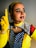 A person is dressed in a pop art-inspired costume featuring bright colors and dotted face paint to mimic a comic book character. The face has red dots and bold black lines emphasizing facial features. The outfit includes a yellow top, gloves, and a cape or scarf with polka dot patterns.