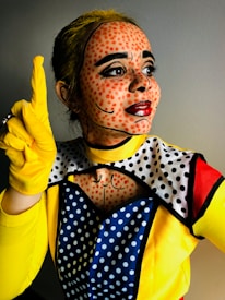 A person is dressed in a pop art-inspired costume featuring bright colors and dotted face paint to mimic a comic book character. The face has red dots and bold black lines emphasizing facial features. The outfit includes a yellow top, gloves, and a cape or scarf with polka dot patterns.