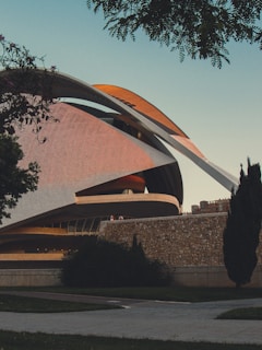 An architecturally striking modern building with sweeping curves and intersecting lines is surrounded by trees and a stone wall. The structure appears to be a large cultural or entertainment venue.