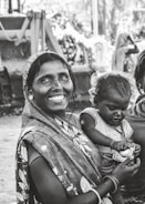A woman dressed in a patterned sari holds a child in her arms, both displaying expressions of joy and contentment. They are positioned outdoors with blurred machinery in the background, suggesting a rural or working-class setting.