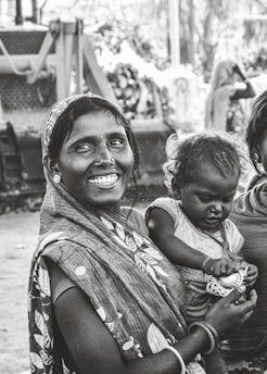 A woman dressed in a patterned sari holds a child in her arms, both displaying expressions of joy and contentment. They are positioned outdoors with blurred machinery in the background, suggesting a rural or working-class setting.