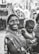 A woman dressed in a patterned sari holds a child in her arms, both displaying expressions of joy and contentment. They are positioned outdoors with blurred machinery in the background, suggesting a rural or working-class setting.