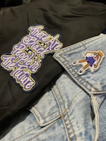 A close-up of clothing featuring a black fabric with an embroidered message in stylized, colorful letters and a light blue denim jacket. The jacket has a patch of a girl in a school uniform.