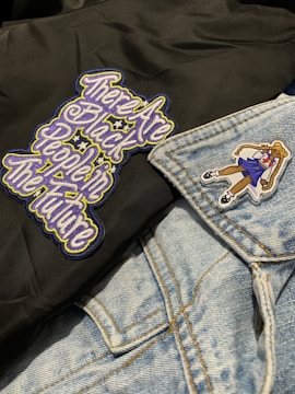 A close-up of clothing featuring a black fabric with an embroidered message in stylized, colorful letters and a light blue denim jacket. The jacket has a patch of a girl in a school uniform.