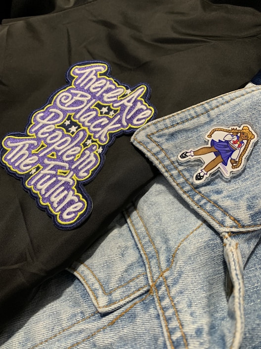 A close-up of clothing featuring a black fabric with an embroidered message in stylized, colorful letters and a light blue denim jacket. The jacket has a patch of a girl in a school uniform.