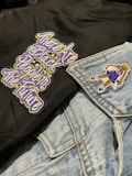 A close-up of clothing featuring a black fabric with an embroidered message in stylized, colorful letters and a light blue denim jacket. The jacket has a patch of a girl in a school uniform.