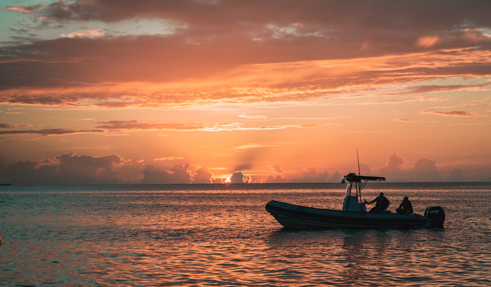 A small boat with passengers silhouetted on calm water under an orange and pink sunset sky