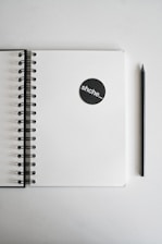 A spiral-bound notebook lies open with a blank page, accompanied by a black pencil positioned parallel to it. On the notebook is a black circular sticker with the text 'shche_'. The background is plain and light in color, giving a minimalistic feel.