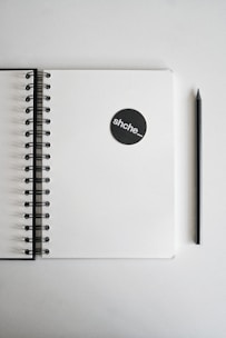 A spiral-bound notebook lies open with a blank page, accompanied by a black pencil positioned parallel to it. On the notebook is a black circular sticker with the text 'shche_'. The background is plain and light in color, giving a minimalistic feel.