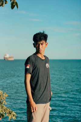 A young man stands near the edge of a body of water under a clear blue sky. He is wearing a dark T-shirt with a small flag patch on the sleeve and light beige pants. A large ship is visible on the calm sea in the background, and a leafy branch can be seen in the upper left corner of the image.