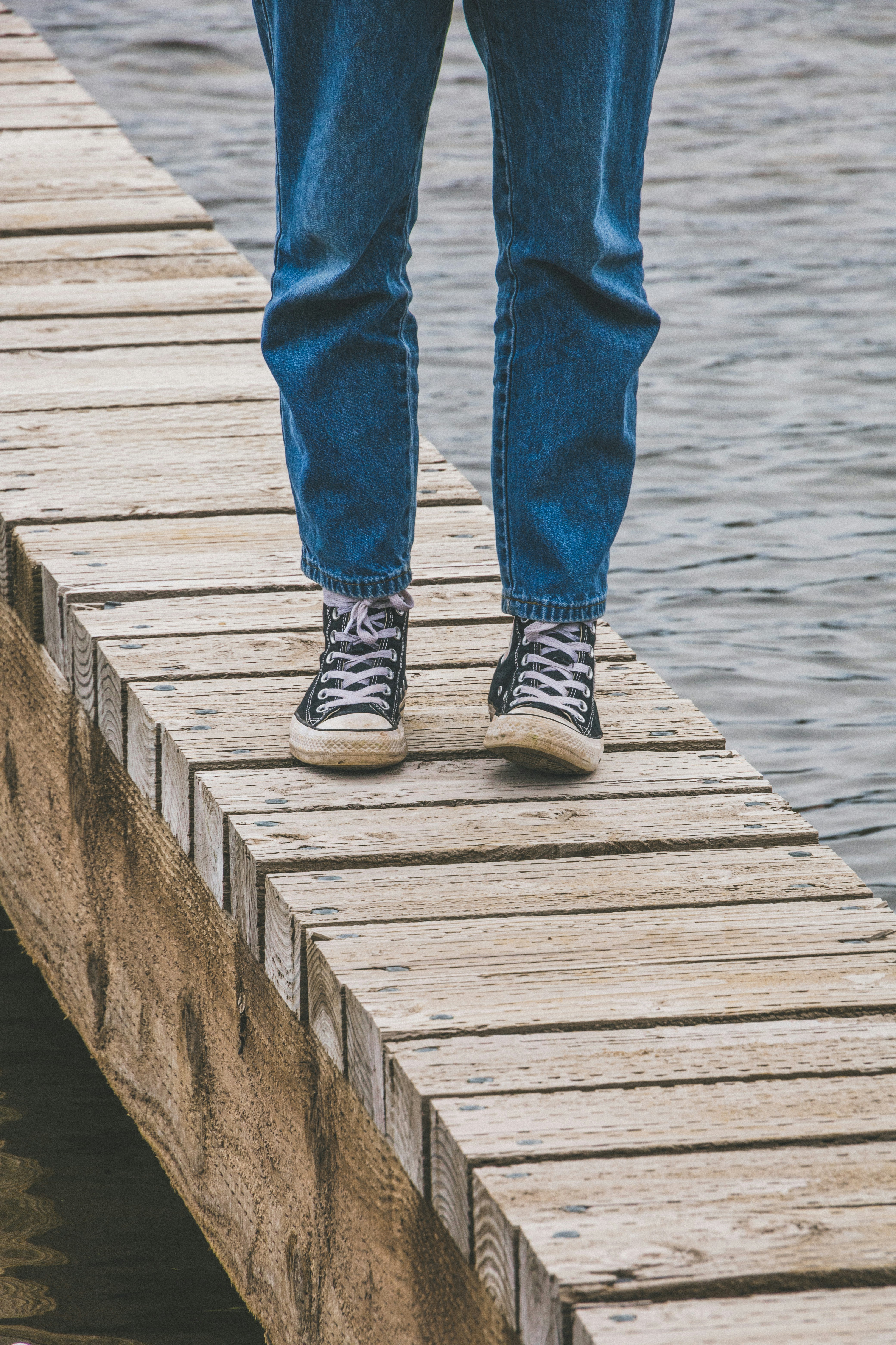 person in blue denim jeans and white and black sneakers standing on wooden dock