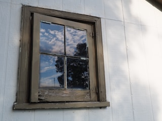A wooden-framed window on a white exterior wall reflects a partly cloudy sky and the outline of nearby trees. The window appears weathered and aged, with signs of peeling paint on the frame.