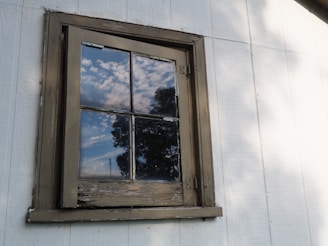 A wooden-framed window on a white exterior wall reflects a partly cloudy sky and the outline of nearby trees. The window appears weathered and aged, with signs of peeling paint on the frame.