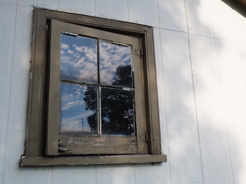 A wooden-framed window on a white exterior wall reflects a partly cloudy sky and the outline of nearby trees. The window appears weathered and aged, with signs of peeling paint on the frame.