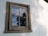 A wooden-framed window on a white exterior wall reflects a partly cloudy sky and the outline of nearby trees. The window appears weathered and aged, with signs of peeling paint on the frame.