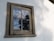 A wooden-framed window on a white exterior wall reflects a partly cloudy sky and the outline of nearby trees. The window appears weathered and aged, with signs of peeling paint on the frame.
