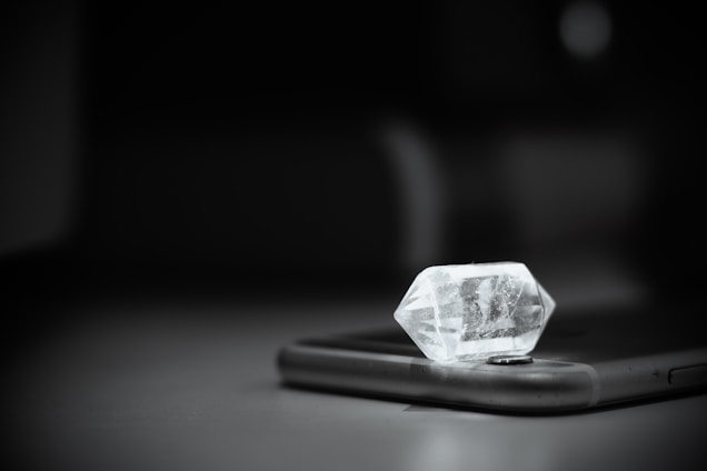 A clear crystal is placed on top of a smartphone, with light reflecting off its faceted surfaces, casting subtle shadows on the device. The background is softly blurred, emphasizing the central objects.