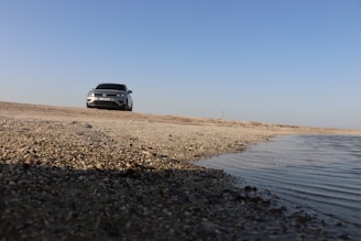 A white car is parked on a sandy coastline with the sea visible on the right side of the image. The sky is clear and blue, and the ground is composed of sand and small rocks.