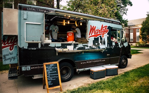 A food truck parked on a paved walkway surrounded by green grass. The truck has a large logo 'Marty's' on its side and is decorated with city skyline graphics. The serving window is open, with two staff members visible inside, and a vertical menu board stands in front. Several coolers are placed on the ground nearby.