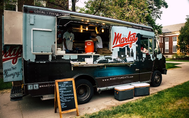 A food truck parked on a paved walkway surrounded by green grass. The truck has a large logo 'Marty's' on its side and is decorated with city skyline graphics. The serving window is open, with two staff members visible inside, and a vertical menu board stands in front. Several coolers are placed on the ground nearby.