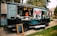 A food truck parked on a paved walkway surrounded by green grass. The truck has a large logo 'Marty's' on its side and is decorated with city skyline graphics. The serving window is open, with two staff members visible inside, and a vertical menu board stands in front. Several coolers are placed on the ground nearby.