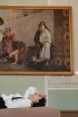 A large, classic painting is mounted on a wall in an ornate setting, with one central figure and several people engaged in various activities. Below the painting, a person is lying on a bench, wearing a white shirt and a black tie, with one arm covering their face.
