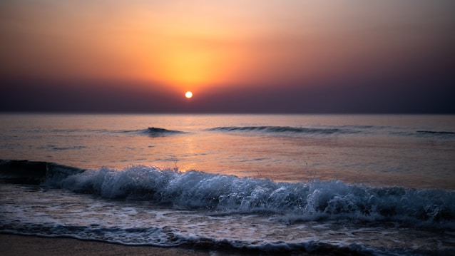 A serene ocean scene with gentle waves lapping at the shore under an early sunrise. The sky transitions from a deep indigo to a warm orange glow, reflecting softly on the tranquil water.