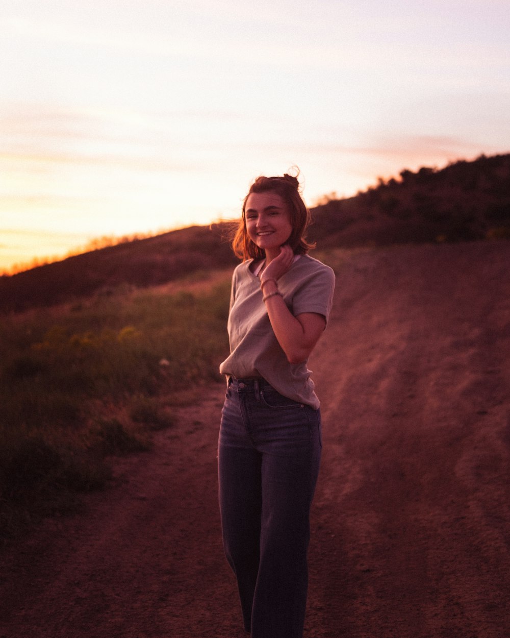 woman in white shirt and blue denim jeans standing on brown dirt road during sunset