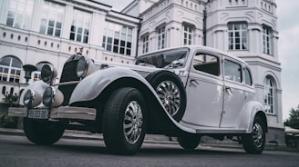 A vintage car is parked in front of a grand, historic building with multiple windows and white decorative elements. The car features a classic design with round headlights and chrome wheels.