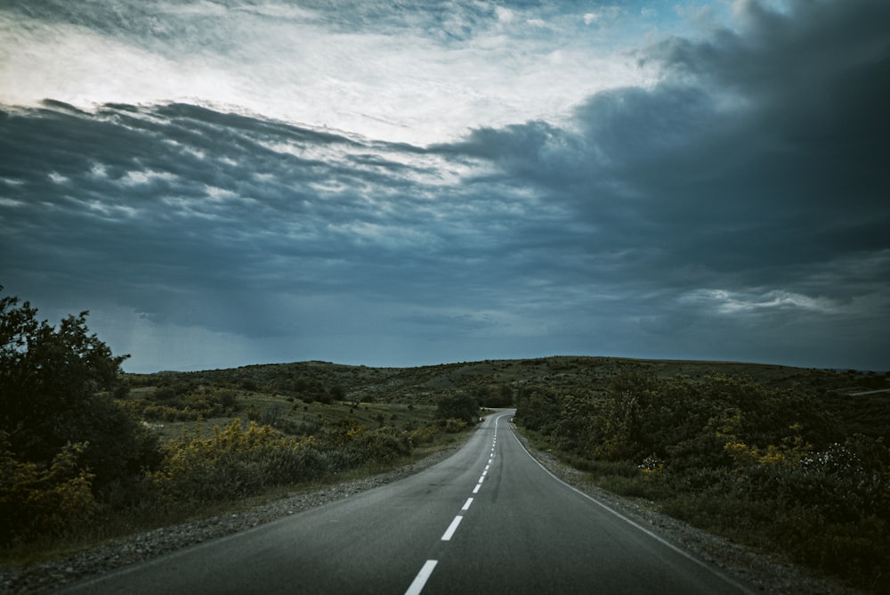 gray asphalt road between green grass field under blue and white sunny cloudy sky during daytime