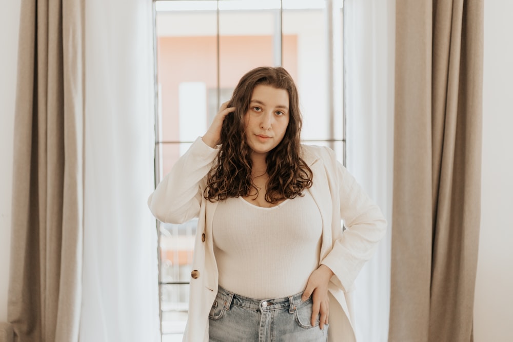 Mujer con crop top blanco de manga larga y jeans de mezclilla azul de pie cerca de la ventana