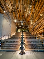 A grand staircase with wooden steps and a unique installation of wooden beams on the ceiling, illuminated by lights running along the center. Metal railings line the sides of the staircase, and the walls have a concrete finish. The setting suggests a modern and artistic interior design.