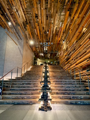 A grand staircase with wooden steps and a unique installation of wooden beams on the ceiling, illuminated by lights running along the center. Metal railings line the sides of the staircase, and the walls have a concrete finish. The setting suggests a modern and artistic interior design.