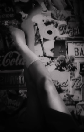 A pair of legs wearing white socks is positioned against a collage-like background featuring various retro elements such as a Coca-Cola sign, a license plate, and the words 'The End'. The image has a vintage and artistic feel, rendered in black and white.