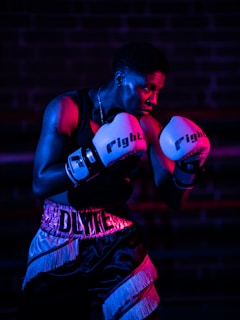 A boxer with a focused expression stands in a fighting stance. They are wearing boxing gloves and shorts, illuminated by dramatic pink and blue lighting against a dark background.