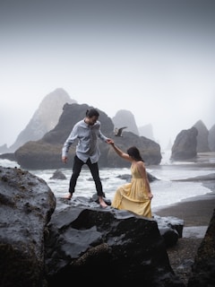 A couple is positioned on large, dark rocks near the sea. The man, in a light shirt and dark pants, is helping the woman, in a flowing yellow dress, over the rocks. Misty rock formations and rough waves create a dramatic coastal background.