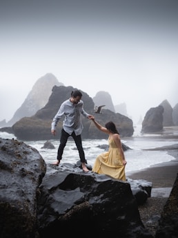 A couple is positioned on large, dark rocks near the sea. The man, in a light shirt and dark pants, is helping the woman, in a flowing yellow dress, over the rocks. Misty rock formations and rough waves create a dramatic coastal background.