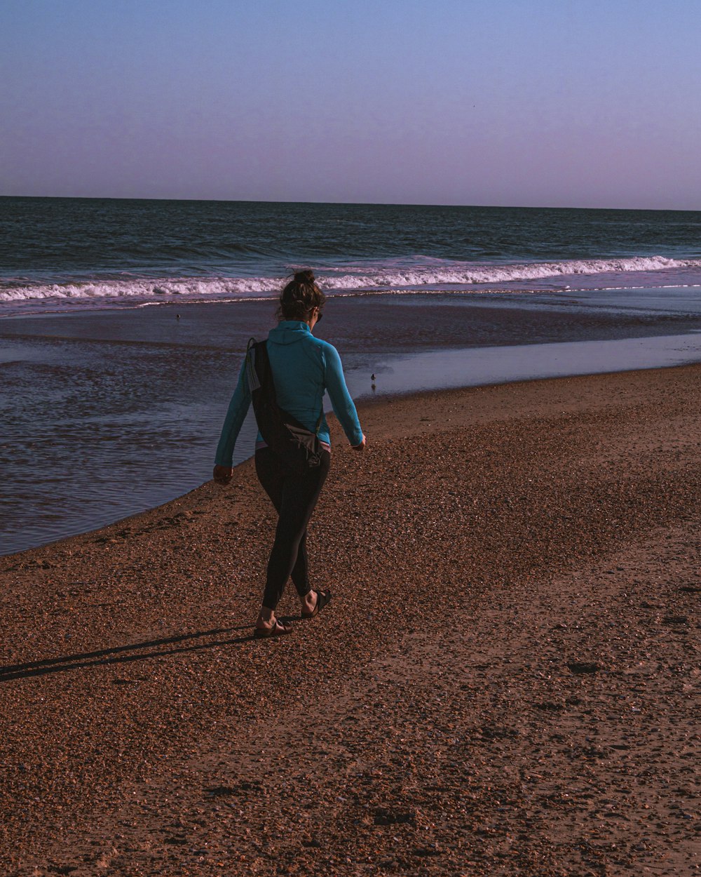 Frau in blauem Langarmhemd und schwarzer Hose geht tagsüber am Strandufer spazieren