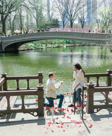 A man is kneeling on a wooden deck next to a body of water, proposing to a woman who looks surprised and delighted. Red rose petals and vases with roses surround them. Behind them, a bridge with the words 'WILL YOU MARRY ME?' in large letters spans the water. Trees and a distant cityscape are visible in the background.