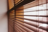 A close-up view of wooden window blinds with light filtering through them, casting shadows and horizontal lines on the surface.