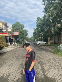 A young person wearing a black and red sports jersey with the number 17 and blue shorts stands on a cobblestone street with a thoughtful expression. The background features various types of greenery, a small commercial building with an Alfamart sign, and parked cars. The sky is mostly clear with some fluffy clouds.