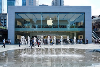 A modern glass-fronted store with a prominent Apple logo, surrounded by skyscrapers. People are visible inside the store and walking in front of it. In the foreground, a fountain with water jets creates a lively atmosphere.