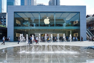 A modern glass-fronted store with a prominent Apple logo, surrounded by skyscrapers. People are visible inside the store and walking in front of it. In the foreground, a fountain with water jets creates a lively atmosphere.